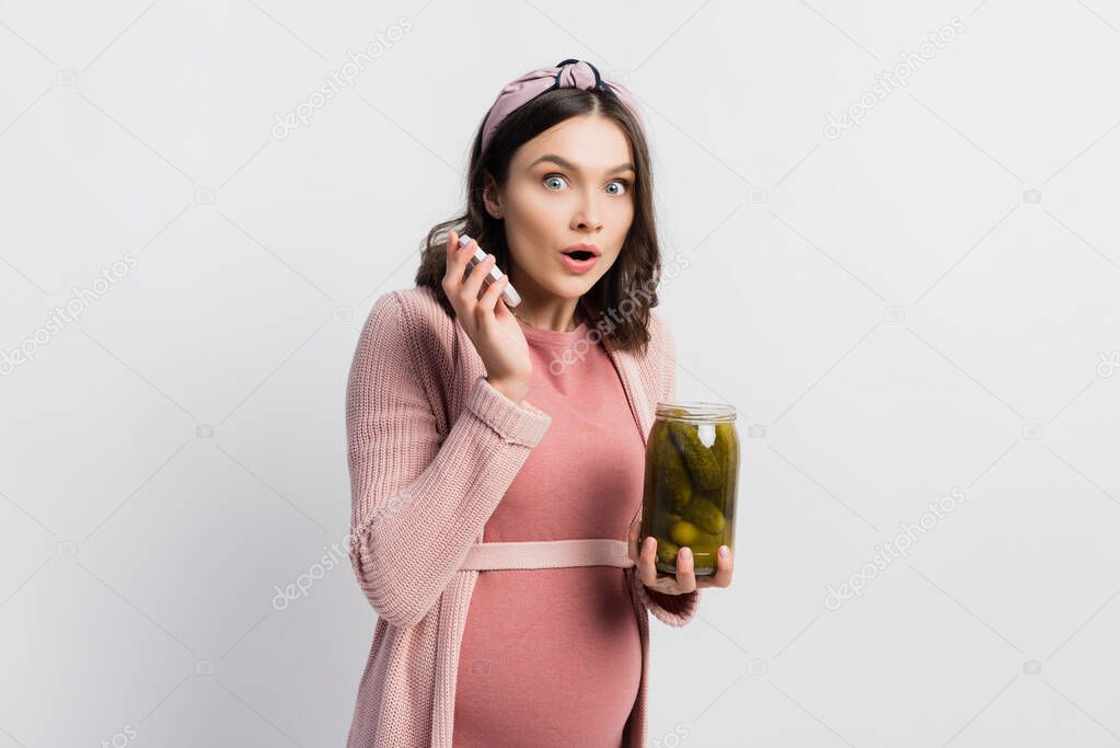 surprised and pregnant woman holding jar with pickled cucumbers on white