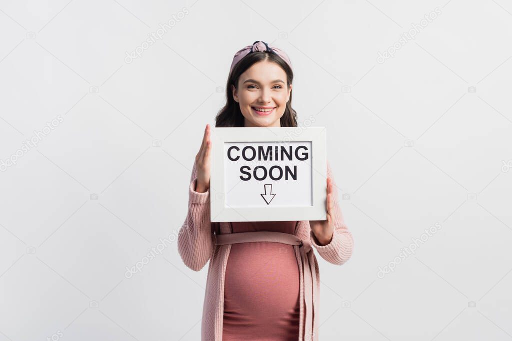 Joyful pregnant woman holding board with coming soon lettering isolated on white