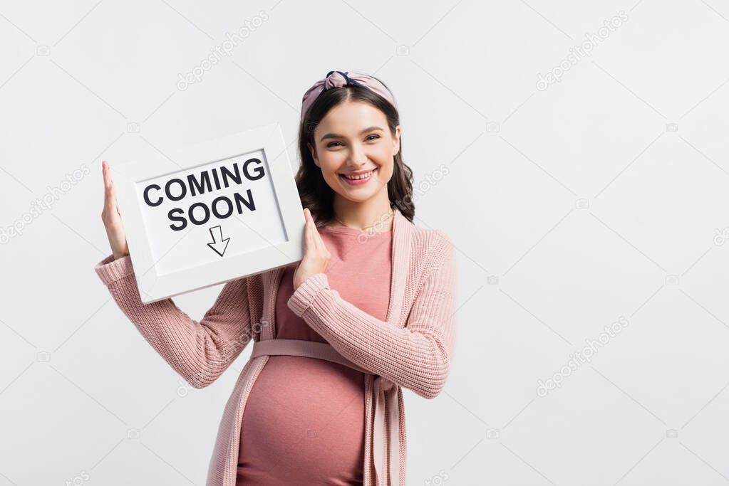 Pleased and pregnant woman holding board with coming soon lettering isolated on white