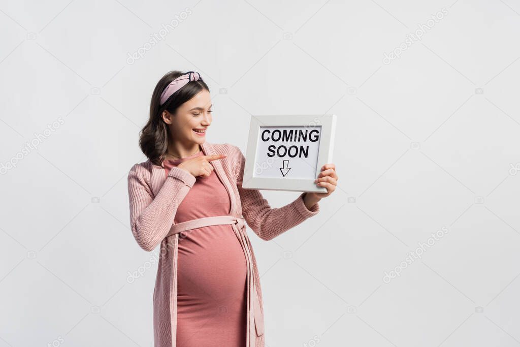 Joyful pregnant woman pointing with finger at board with coming soon lettering isolated on white