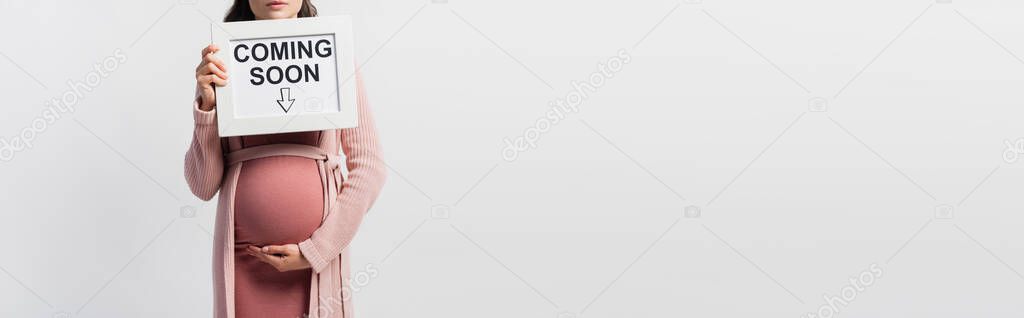 Panoramic crop of pregnant woman holding board with coming soon lettering isolated on white