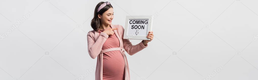 Joyful pregnant woman pointing with finger at board with coming soon lettering isolated on white