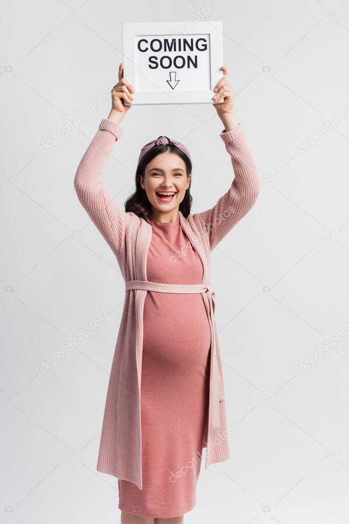 Excited and pregnant woman holding board with coming soon lettering isolated on white