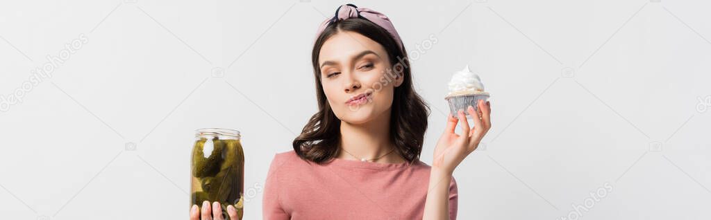 panoramic shot of woman choosing between cupcake and jar with pickled cucumbers isolated on white