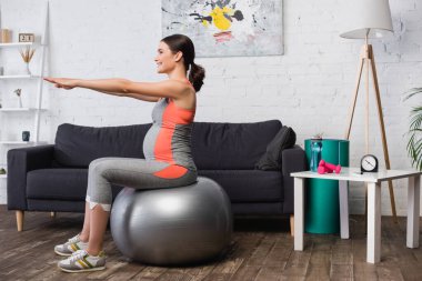 Spor kıyafetli hamile bir kadın oturma odasında spor paspasıyla egzersiz yapıyor. 