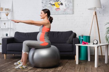 pleased pregnant woman in sportswear exercising on fitness mat in living room  clipart