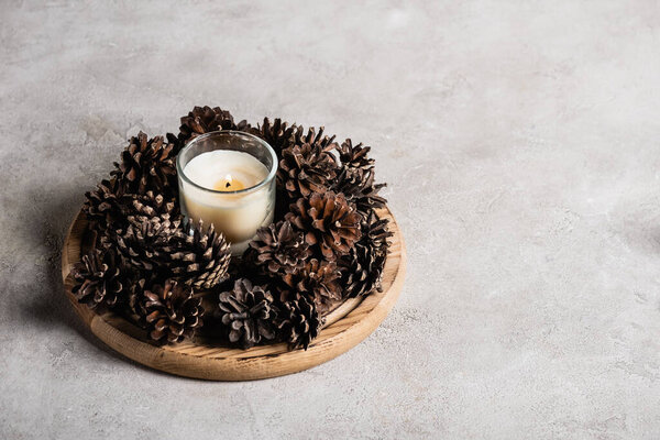 Scented candle with pine cones on wooden plate on grey and textured background