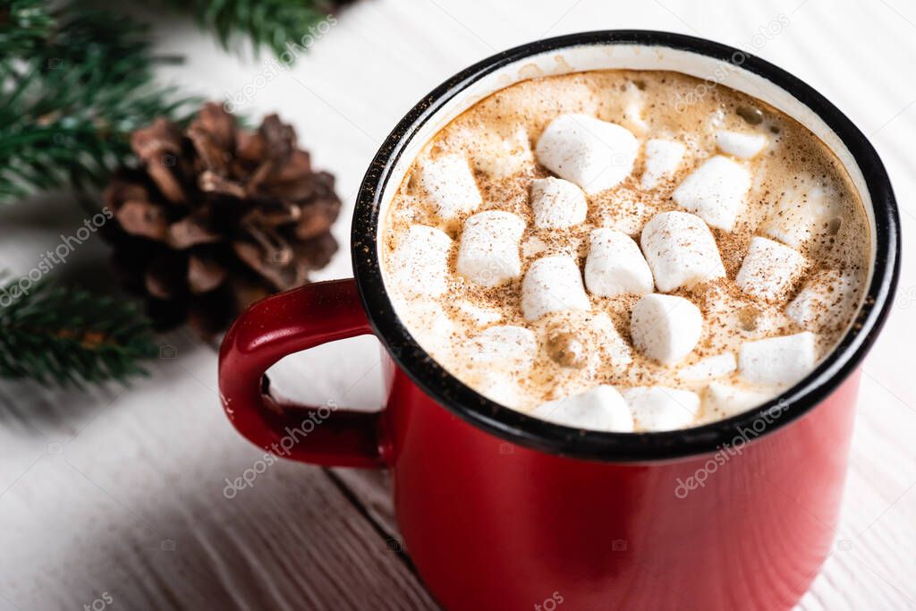 Close up view of red cup of cocoa with marshmallows and cinnamon near pine cone