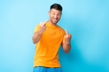 smiling man showing middle fingers and looking at camera on blue  clipart