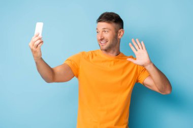 man in t-shirt holding smartphone and taking selfie while waving hand on blue clipart