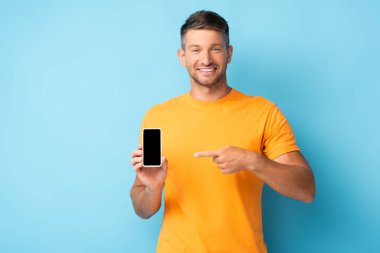 Tişörtlü mutlu adam mavi ekranlı akıllı telefonu işaret ediyor.