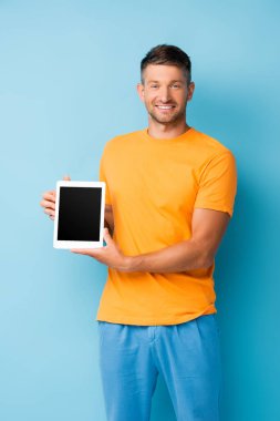 Tişörtlü memnun bir adam elinde mavi ekranlı dijital tablet tutuyor.