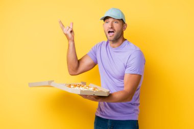 Mavi şapkalı bir adam elinde lezzetli pizzayla kutuda el kol hareketi yapıyor. 