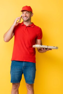 Kırmızı şapkalı mutlu teslimatçı elinde lezzetli pizzayla sarıda akıllı telefondan konuşuyor.