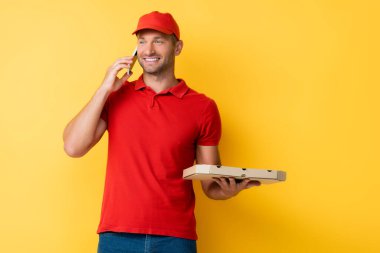 Kırmızı şapkalı gülümseyen teslimatçı elinde lezzetli pizzayla sarıda akıllı telefondan konuşuyor.