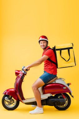 Kasklı ve sırt çantalı mutlu teslimatçı sarı mobiletle kırmızı scooter sürüyor.