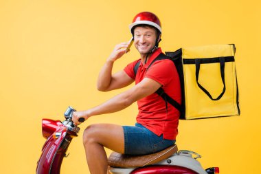 Mutlu teslimatçı kasklı ve sırt çantalı kırmızı scooter 'la sarı telefonda konuşurken.
