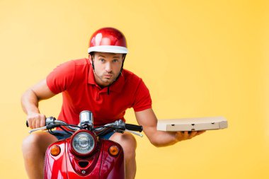 concentrated delivery man in helmet riding red scooter and holding pizza box isolated on yellow clipart