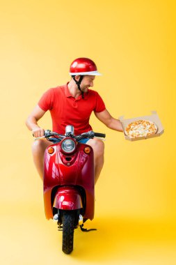 Kırmızı scooter süren kasklı gülümseyen teslimatçı ve sarı kutuda pizza tutan.