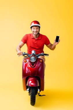 Kasklı mutlu teslimatçı scooter sürüyor ve elinde sarı ekranlı akıllı telefon tutuyor. 