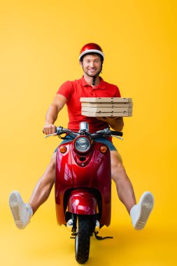 Kasklı gülümseyen teslimatçı scooter sürüyor ve sarı kutular üzerinde pizza kutuları taşıyor. 