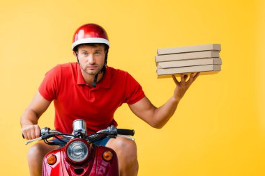 Kasklı ciddi bir teslimatçı scooter sürüyor ve sarı kutular üzerinde pizza kutuları taşıyor. 