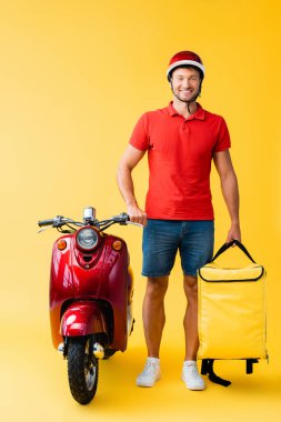 Sırt çantalı neşeli teslimatçı kırmızı scooter 'ın yanında duruyor.