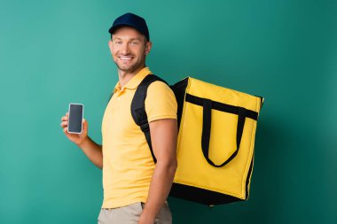 Sarı sırt çantası taşıyan ve elinde mavi ekranlı akıllı telefon tutan neşeli teslimatçı.