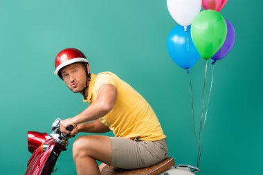 Kasklı teslimatçı elinde balonlarla kırmızı scooter sürüyor.