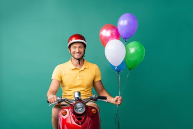 Kırmızı scooter kullanan kasklı gülümseyen teslimatçı mavi üzerinde balonlar tutarken