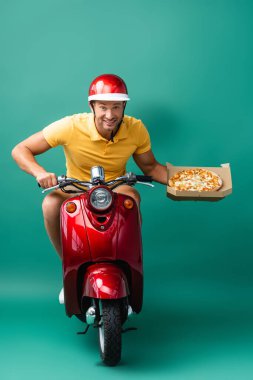 Kasklı neşeli teslimatçı elinde lezzetli pizzayı mavi kutuda tutarken scooter kullanıyor.