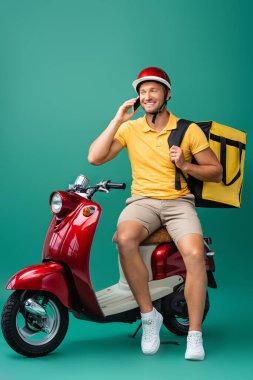 Sırt çantalı neşeli teslimatçı, mavi motosikletin yanında akıllı telefondan konuşuyor.