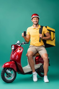 Sırt çantalı neşeli teslimatçı elinde mavi motosikletin yanında boş ekranlı akıllı telefon tutuyor.