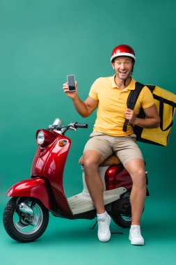 Sırt çantalı heyecanlı teslimatçı elinde mavi motosikletin yanında boş ekranlı akıllı telefon tutuyor.