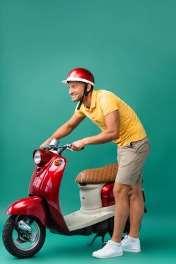 Kırmızı scooter 'ın yanında duran kasklı mutlu teslimatçı.