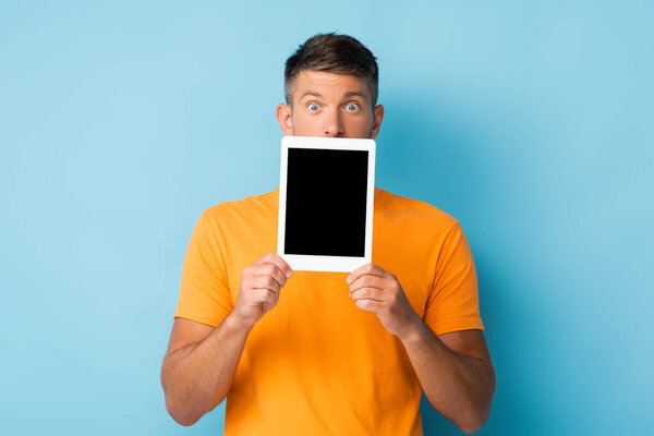 shocked man in t-shirt covering mouth while holding digital tablet with blank screen on blue