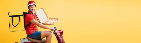 happy delivery man with backpack riding red scooter and holding pizza in carton box on yellow, banner