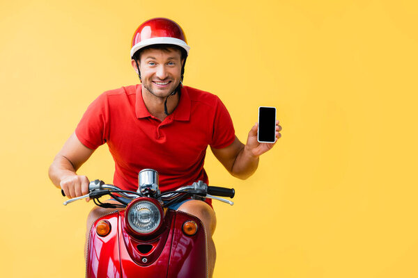 pleased delivery man in helmet riding scooter and holding smartphone with blank screen on yellow 