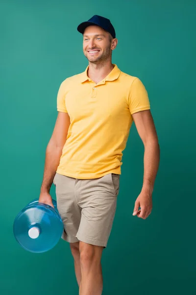 cheerful delivery man in cap carrying bottle of water on blue