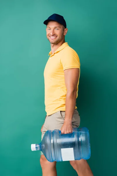 smiling delivery man in cap carrying bottle of water on blue