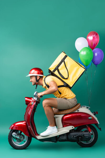 side view of focused delivery man with backpack riding scooter with balloons on blue 