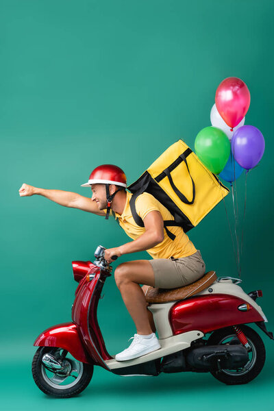side view of happy delivery man with backpack and outstretched hand riding scooter with balloons on blue 