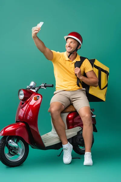 cheerful deliver man with backpack taking selfie near scooter on blue