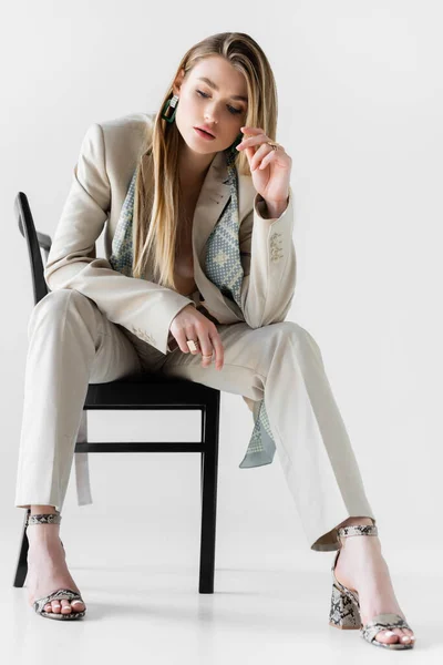 trendy woman in suit sitting on chair and looking down on white