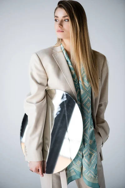 stylish woman in suit holding round mirror while looking away on grey