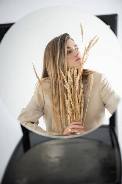 reflection of young woman looking away while holding wheat with blurred chair on background