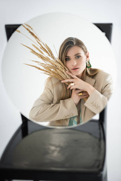 reflection of young woman looking at camera while holding wheat with blurred chair on background