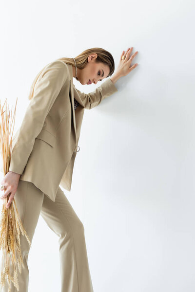 young woman in beige formal wear holding wheat and leaning on white wall