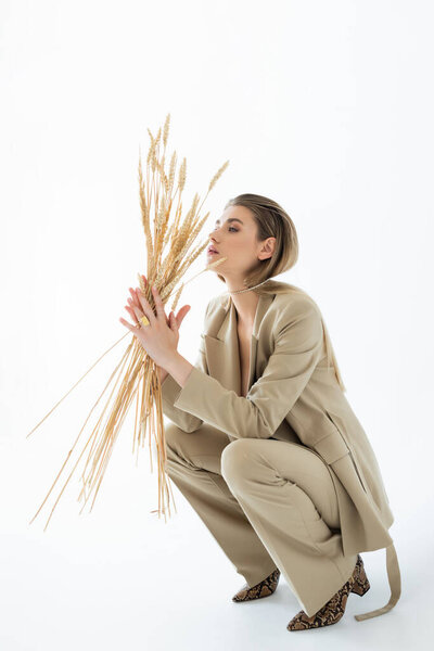 young woman in beige suit sitting while holding wheat on white