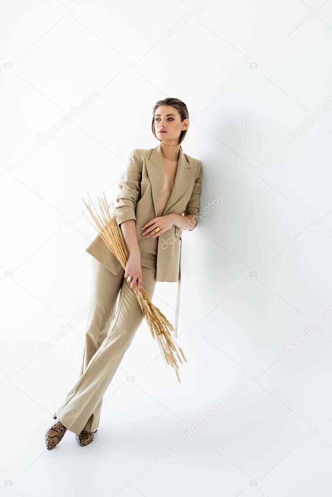 full length of young woman in beige formal wear posing while holding wheat on white